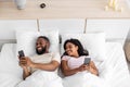 Happy laughing young african american wife and husband with smartphones lies on comfortable bed in bedroom at free time Royalty Free Stock Photo