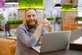 Happy laughing young handsome bearded trendy man sitting with silver laptop in cafe during coffee break Royalty Free Stock Photo