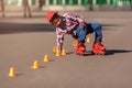 Happy little african american boy on rollers skates in summer park at asphalt road among training cones . Roller kid