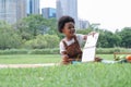 Happy little African kid boy holding a brush to paint in book while sitting on picnic mat in park Royalty Free Stock Photo