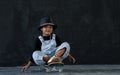 Happy little African kid boy wear hat, necklace chain and jeans bib practice playing and sitting on skateboard pushing cement Royalty Free Stock Photo