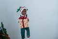 Happy little African kid girl with green bib clothes and reindeer Santa hat standing and holding gift box in Xmas holiday at home