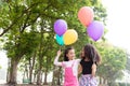 Happy little asian & african american girl playing colorful balloons together. Trees and green gardens background. Smiling lovely Royalty Free Stock Photo