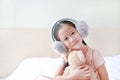 Happy little Asian child girl wearing winter earmuffs and embracing teddy bear while sitting on the bed at home Royalty Free Stock Photo