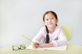 Happy little Asian child girl write in a book or notebook with pencil sitting on kid chair and table against white background Royalty Free Stock Photo