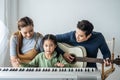 Happy little Asian daughter playing piano with mother and father play guitar at home, Mother teaching daughter to play piano, Royalty Free Stock Photo