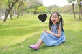 Happy little Asian kid girl holding blank heart label sitting on green grass at garden outdoor Royalty Free Stock Photo