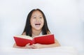 Happy little Asian kid girl reading hardcover book lying with pillow on bed against white background Royalty Free Stock Photo