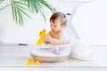 Happy little baby girl washes in a basin with foam and water in a bright room at home and plays with a yellow rubber duck Royalty Free Stock Photo