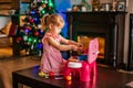 Happy little blonde girl playing near Christmas tree with toy kitchen. Xmas morning in decorated living room with fireplace and