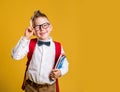 Happy little boy in glasses with books and bag on his first day to school. Funny child against yellow background Royalty Free Stock Photo