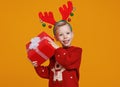 Happy little boy in red Christmas sweater and deer antlers holding Xmas gift against yellow wall Royalty Free Stock Photo