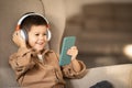Happy little boy in wireless headphones watch video on smartphone, sits in armchair, calls on phone