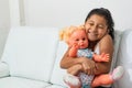 Happy little brunette girl hugging her doll tightly ready to play while sitting on the white sofa Royalty Free Stock Photo