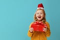 Happy little child girl in a warm knitted sweater and cap Santa Claus is holding a red box with a gift on a blue background. Royalty Free Stock Photo