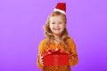 Happy little child girl in a warm knitted sweater and cap Santa Claus is holding a red box with a gift on a purple background. Royalty Free Stock Photo