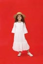 Happy little curly girl, wear in white dress and hat, looking at camera, on red background. Vertical view. Royalty Free Stock Photo