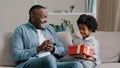 Happy little daughter sitting on sofa with closed eyes receiving gift box from loving father caring dad congratulating Royalty Free Stock Photo