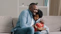 Happy little daughter sitting on sofa with closed eyes receiving gift box from loving father caring dad congratulating Royalty Free Stock Photo