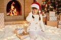 Happy little girl in beautiful white sweater and red santa hat in front of New Year Christmas tree, smiling, poses and holding Royalty Free Stock Photo