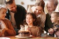 Happy little girl celebrating birthday with family at home, closing eyes and making wish with cake with lit candles