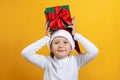 Happy little girl with christmas present on yellow background. A child in a hat holds a gift on his head Royalty Free Stock Photo