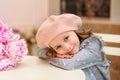 Happy little girl with curly hair in a dress and in a beret sits at a table with a bouquet of pink peonies in a street vintage caf Royalty Free Stock Photo