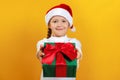 Happy little girl with a gift for Christmas. A child in a Christmas hat gives a gift on a yellow background Royalty Free Stock Photo