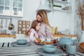 Happy little girl and her beautiful young mother have breakfast together in a white kitchen. Mom hugs and kisses daughter. Royalty Free Stock Photo