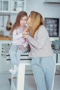 Happy little girl and her beautiful young mother have breakfast together in a white kitchen. Mom hugs and kisses daughter. Royalty Free Stock Photo