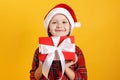 Happy little girl holding a box with a christmas gift. Baby in a red dress and santa hat on a yellow background