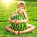 Happy little girl is hugging huge watermelon sitting on the green grass in summertime.