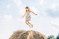 Happy little girl jump up at top of haystack with hands to sides and look down. Have fun on hayrick. Beautiful blue sky.