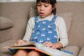Happy little girl reading a book indoors. Cute little girl in casual clothes sitting on the sofa reading a book Royalty Free Stock Photo