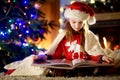 Happy little girl reading a story book by a fireplace in a cozy dark living room on Christmas eve