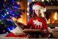 Happy little girl reading a story book by a fireplace in a cozy dark living room on Christmas eve