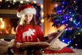 Happy little girl reading a story book by a fireplace in a cozy dark living room on Christmas eve