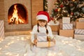 Happy little girl in Santa hat holding Christmas gift boxes while sitting on floor, child wearing white jumper and red santa claus Royalty Free Stock Photo