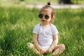 Happy little girl sits on the grass outdoors. Cute baby girl in white bodysuit and sunglasses on the backyard. Adorable Royalty Free Stock Photo