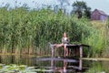 Happy little girl sitting on old wooden jetty surrounded by high green reeds splashing water with her feet at warm summer day. Royalty Free Stock Photo