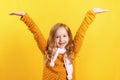 Happy little girl in a sweater and scarf on a yellow background. The child raised his hands up. Concept for autumn, holiday, sale.