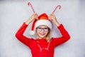 Happy little girl wearing glasses and Santa hat and holding candy canes by her head to make reindeer antlers