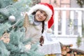 A happy little girl in a white fur coat and santa hat peeks out from behind a Christmas tree against the backdrop of a New Year`s Royalty Free Stock Photo