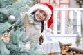 A happy little girl in a white fur coat and santa hat peeks out from behind a Christmas tree against the backdrop of a New Year`s Royalty Free Stock Photo