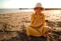 Happy little girl in yellow cute dress and summer hat enjoying sunny day at the beach, sitting on the sand and smiling to the Royalty Free Stock Photo