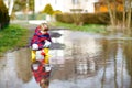 Happy little kid boy in yellow rain boots playing with paper ship boat by huge puddle on spring or autumn day. Active Royalty Free Stock Photo