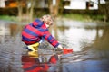 Happy little kid boy in yellow rain boots playing with paper ship boat by huge puddle on spring or autumn day. Active Royalty Free Stock Photo