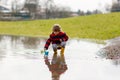 Happy little kid boy in yellow rain boots playing with paper ship boat by huge puddle on spring or autumn day. Active Royalty Free Stock Photo