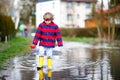 Happy little kid boy in yellow rain boots playing with paper ship boat by huge puddle on spring or autumn day. Active Royalty Free Stock Photo