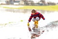 Happy little kid boy in yellow rain boots playing with paper ship boat by huge puddle on spring or autumn day. Active Royalty Free Stock Photo